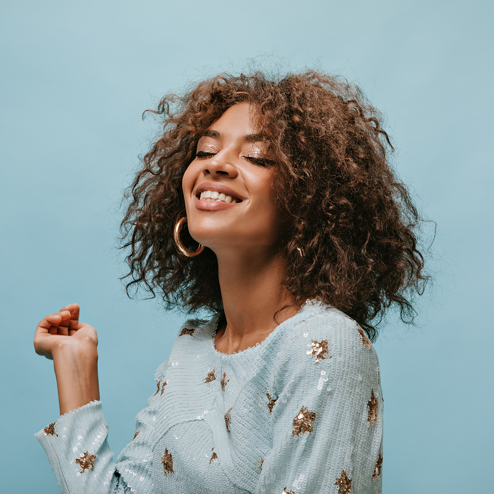 Charming woman with brunette short hair in stylish gold earrings and printed blue clothes smiling with closed eyes on isolated backdrop..